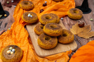 Kürbis Donuts auf einem Holzbrett. Im Hintergrund Hexenhüte, Kastanien und Blätter.